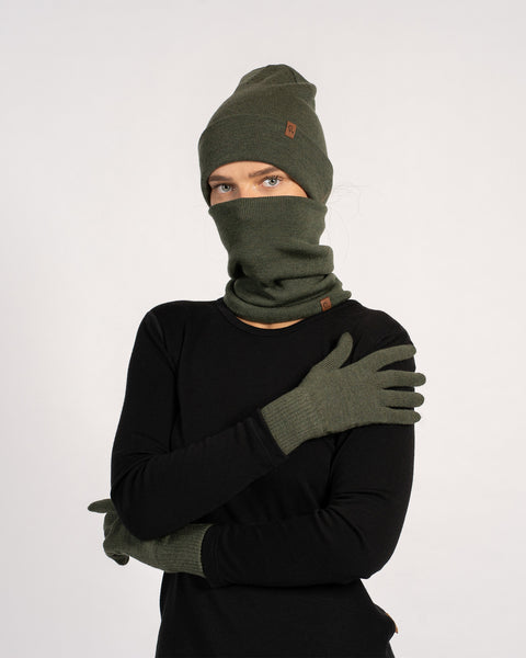 Woman wearing a matching dark green winter set including a beanie, neck warmer pulled up over the face, and gloves, dressed in a black long-sleeve top, posing with arms crossed against a plain light background.