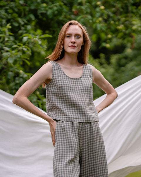 The model is wearing the green gingham crop top ANA. She is facing the camera, in the background there is a white sheet visible. She has both of her arms on the sides of her hips.