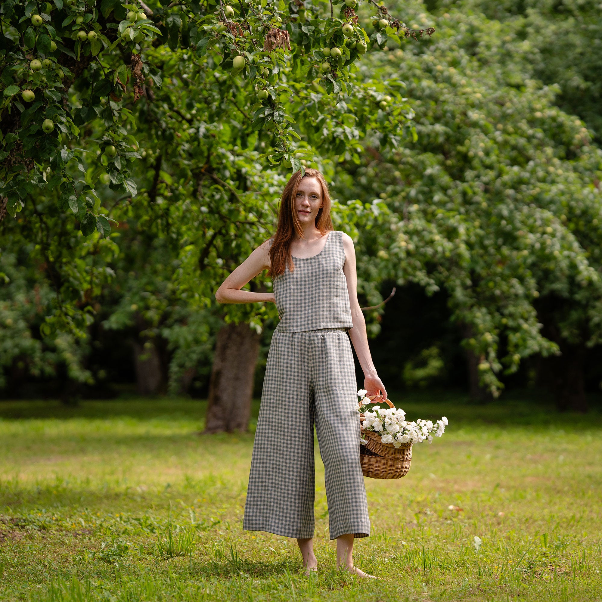 The model is posing sideways, revealing the elastic band at the back of her green gingham pattern linen pants. It is visible, that the pants are slightly cropped and a bit above her ankles. She is holding a basket full of flowers. 
