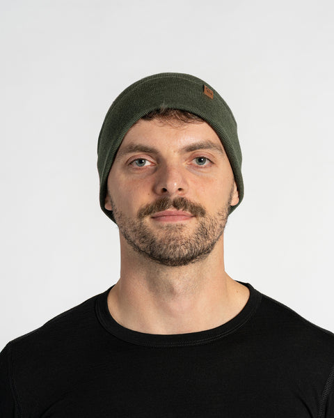A man with light skin, blue eyes, and a beard is looking forward. He wears a dark green colored menique knit beanie with a small brown patch on the side, and a black crew-neck shirt.