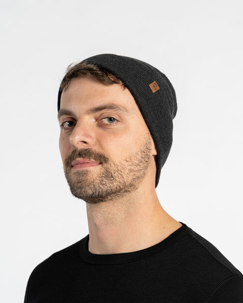 A man with light skin, blue eyes, and a beard smiles faintly while looking forward. He wears a dark gray colored menique knit beanie with a small brown patch on the side, and a black crew-neck shirt.