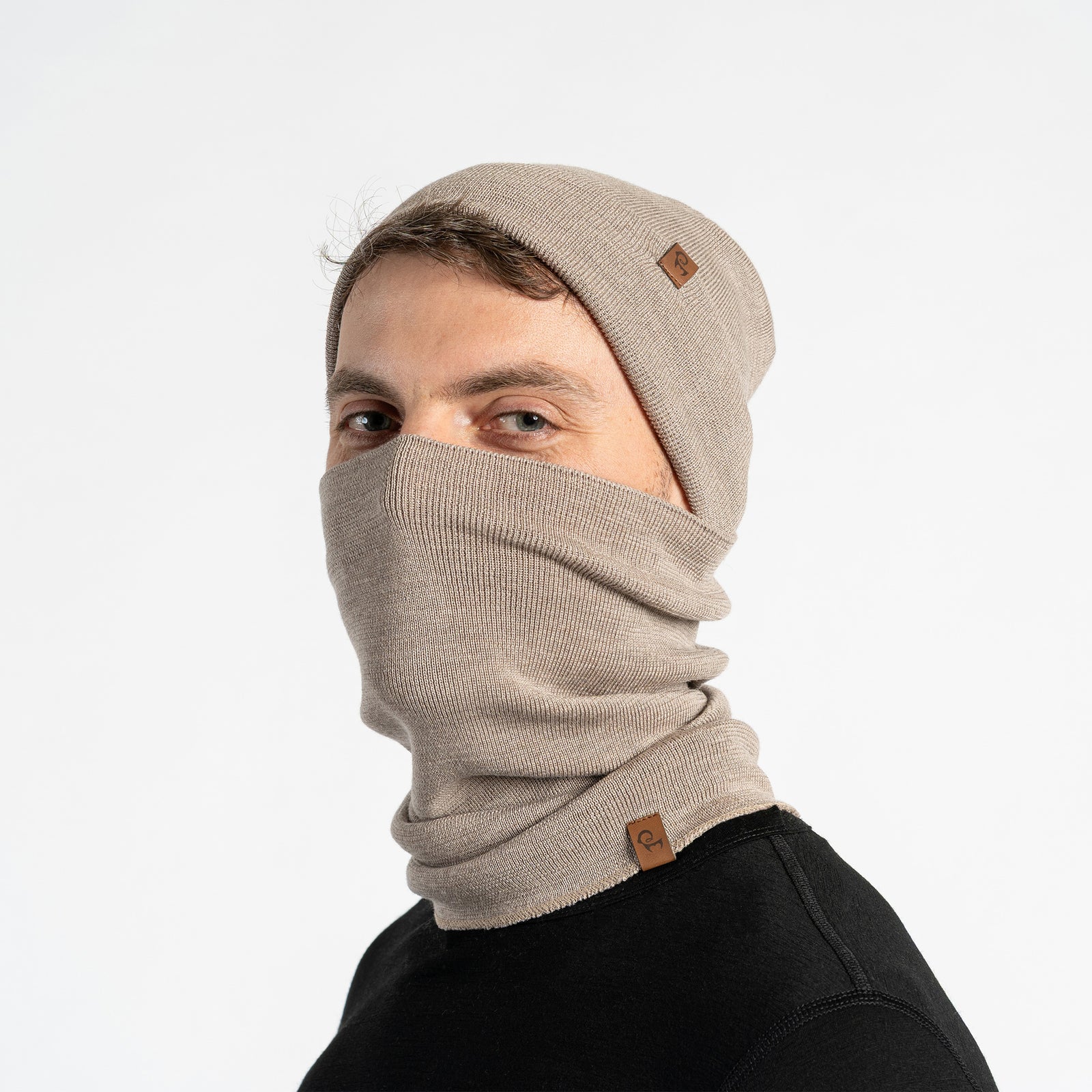 A smiling man with short dark hair and a beard, wearing a creamy beige knit neck warmer and a black long-sleeved shirt, looks directly at the camera. A small brown leather tag is visible on the front of the neck warmer. The background is plain white.