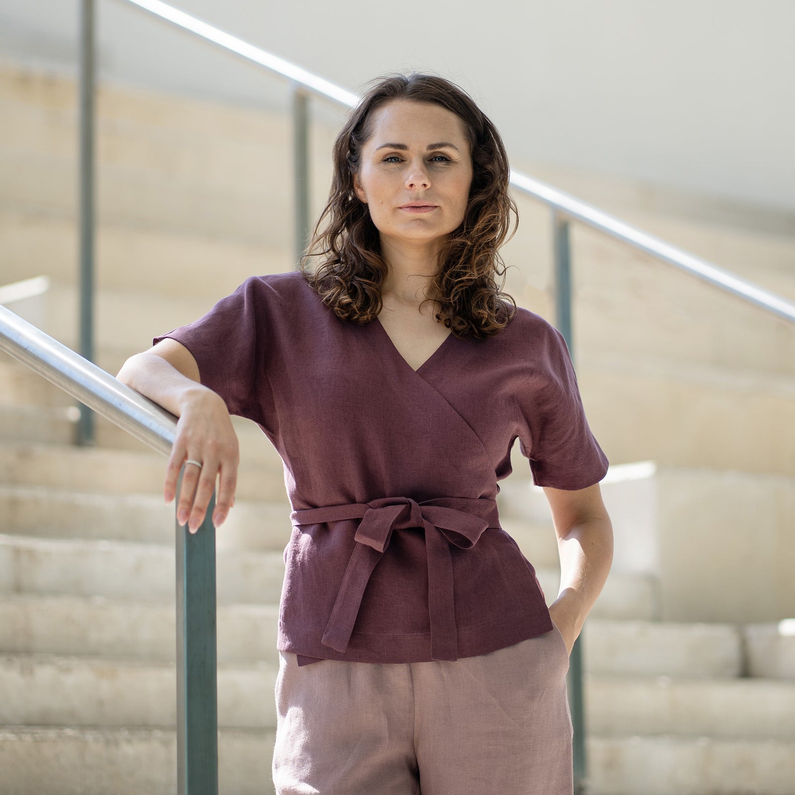A woman with brown hair, wearing a dark purple wrap top and light mauve trousers, smiles while posing on indoor concrete stairs.