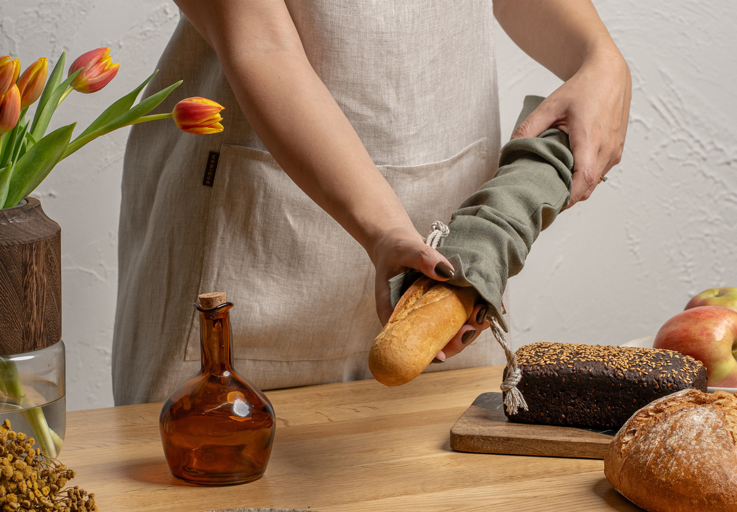 A person in a beige linen apron from menique places a baguette into a green linen bread bag on a wooden table, surrounded by artisanal loaves, apples, tulips, and a glass bottle.