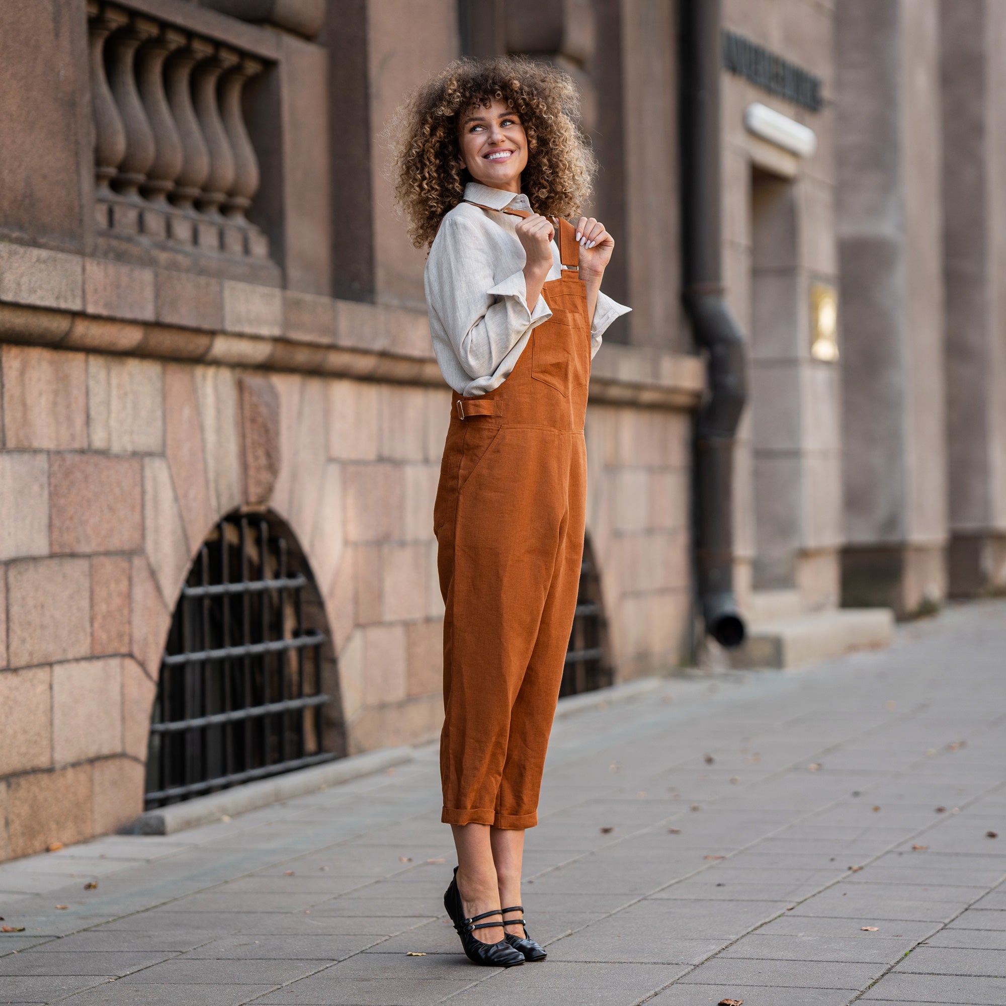 Woman with curly hair standing in a street and wearing Linen Pinafore Jumpsuit Nicci Almond Brown