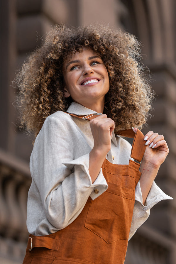 Woman with curly hair standing in a street and wearing Linen Pinafore Jumpsuit Nicci Almond Brown