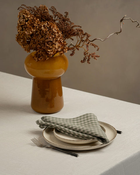 A tabletop covered with a linen tablecloth, with a vase and two stacked plates on top of it. There is a green check napkin on top of the plates.