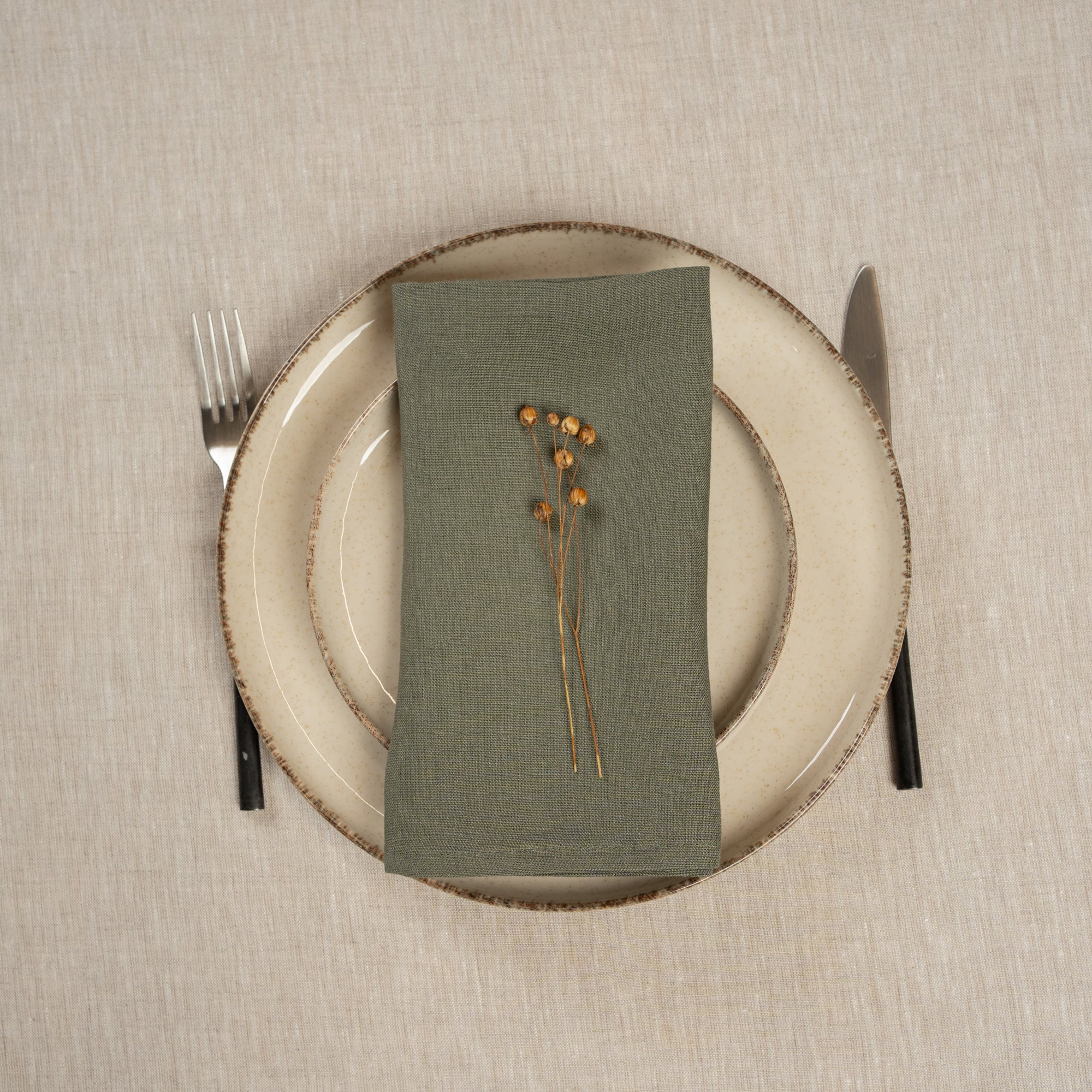 The menique linen napkin in stone green color, put on top of two stacked plates . There are cutlery items by the sides of the plates. The background is a natural color linen tablecloth.