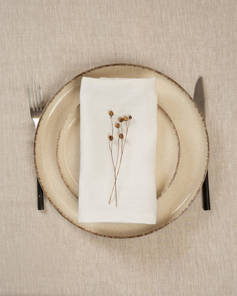 The menique linen napkin in pure white color, put on top of two stacked plates . There are cutlery items by the sides of the plates. The background is a natural color linen tablecloth.