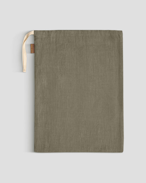 A menique stone green linen storage bag lies flat on a light gray surface, its light-colored drawstrings gathered at the top left, next to a small brown tag.