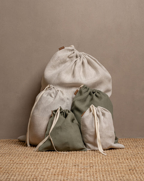 Five closed linen drawstring bags in varying sizes and colors of natural and stone green are arranged on a textured jute rug against a warm brown wall.