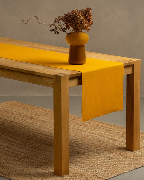 A wooden table positioned on top of a carpet. There is a spicy yellow linen table runner and a vase on top.
