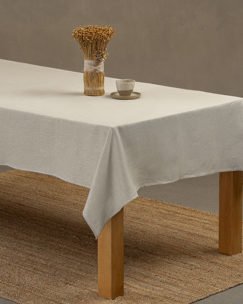 The menique linen tablecloth in pure white color, draping over a wooden table, with a vase and a clay cup and plate set on top of it.