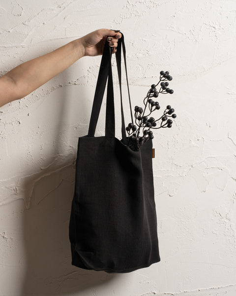 A fair-skinned arm and hand hold up a black reusable linen tote bag against a textured off-white wall. Dark, berry-like stems are visible inside the bag.