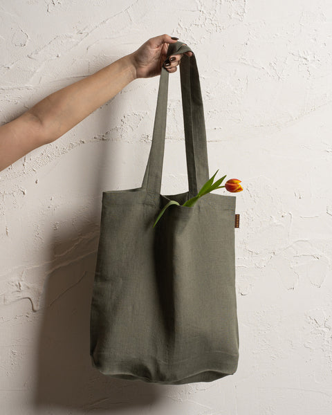 A fair-skinned arm and hand hold up a stone green reusable linen tote bag against a textured off-white wall. A single tulip with a red and yellow bloom and green leaves is visible inside the bag.