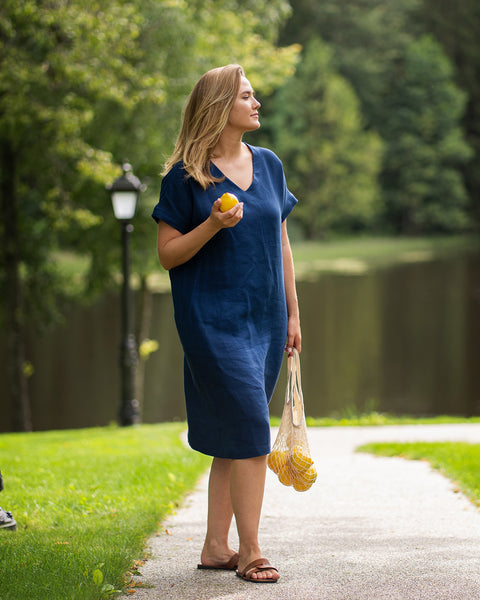 A woman strolls along a park path in a storm blue linen dress and brown slip-on sandals, looking upward into the distance. She holds a single lemon in one hand and carries a string bag filled with more lemons in the other.