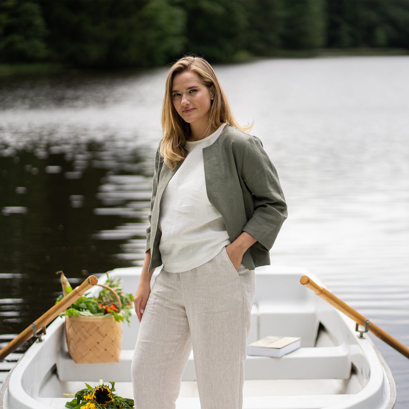 A smiling woman relaxes in a white rowboat on the water, resting her cheek gently against her hand. She is wearing a collarless stone green linen jacket, a white top, and natural linen pants, with a bunch of bright sunflowers resting on her lap.