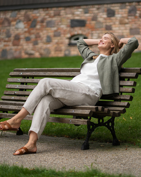A woman relaxes on a wooden park bench, leaning back comfortably with her hands behind her head and her eyes closed in the sunlight. She is wearing a stone green linen jacket over a pure white top, paired with natural-colored linen trousers and brown woven sandals.