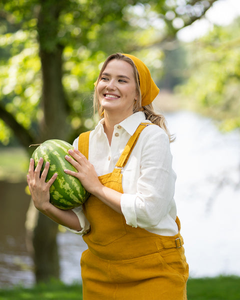 Woman in the mustard yellow outfit stands in a sunlit park with trees and a body of water in the background. She is smiling brightly while holding a large, whole watermelon in her arms.