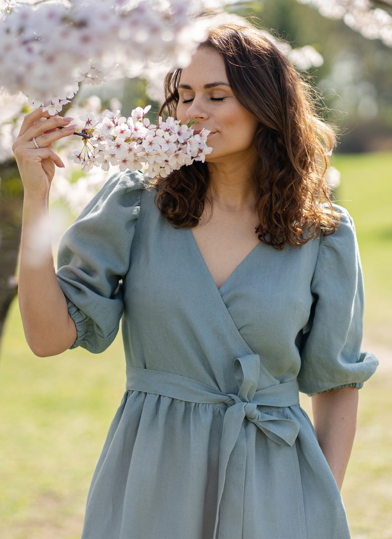 Woman wearing light green linen wrap dress smelling spring blossom tree outdoors, breathable women’s linen dress designed for warm weather, natural fabric clothing for spring and summer.