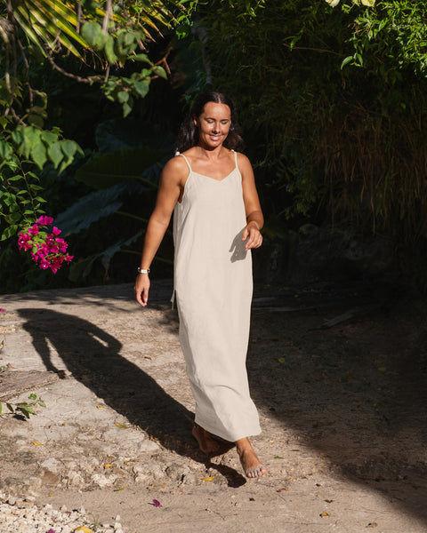 A woman with dark hair wearing a natural linen sundress. She is surrounded by palm trees and tropical foliage. The ground is light-colored with shadows.