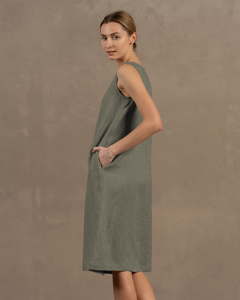 A woman with light skin and her hair styled back is shown in profile, facing slightly away from the viewer, wearing a sleeveless, stone green linen dress and resting one hand in the dress&#39;s side pocket.