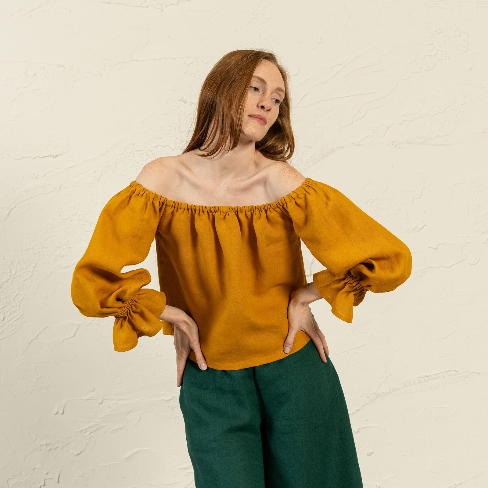 A light-skinned woman with long, straight auburn hair stands in front of a textured light-colored wall, looking slightly to her left. She is wearing a yellow off-the-shoulder top with voluminous sleeves gathered at the wrists and dark green pants. Her hands are on her hips.