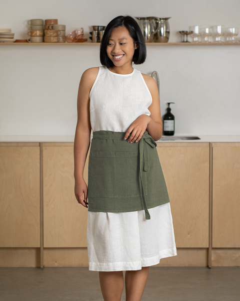 Woman wearing a stone green linen half apron over a white dress, standing in a minimalist kitchen