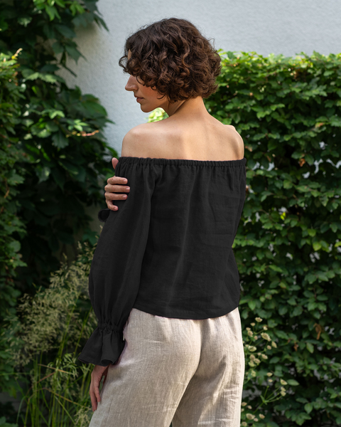 A woman with short, curly brown hair stands with her back mostly to the viewer, looking over her left shoulder, against a backdrop of a dense green ivy wall. She is wearing a black off-the-shoulder top with full sleeves that gather at the wrists and light-colored linen pants. Her left arm is slightly raised with her hand near her shoulder.