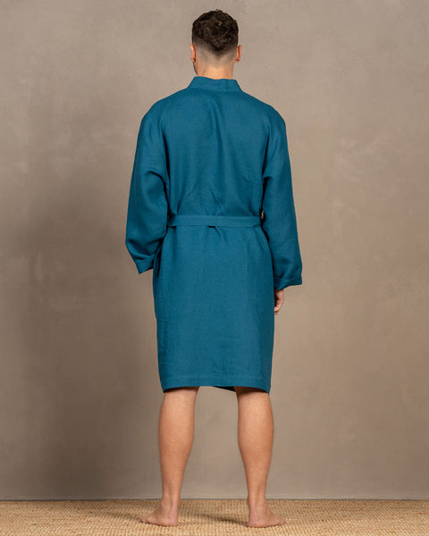 A man standing barefoot on a rug, wearing the menique cobalt blue men&#39;s linen bathrobe. There is a visible tie string at the back. The robe is knee-length.