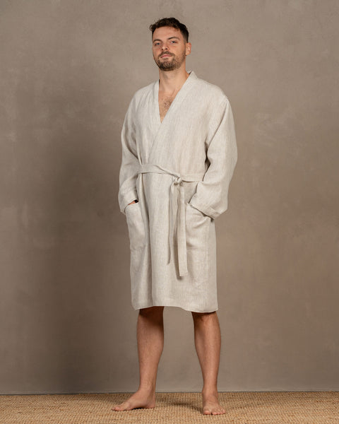 A man, looking at the camera, wearing the menique men&#39;s linen bathrobe in natural color, with both of his hands inside the side pockets. The front tie is tied into a loose knot, the robe features a v-neck cut at the front. The bathrobe is knee-length.