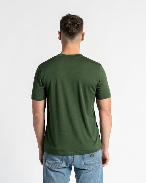 Back view of a man in a merino wool dark green t-shirt and light blue jeans, standing against a white background.