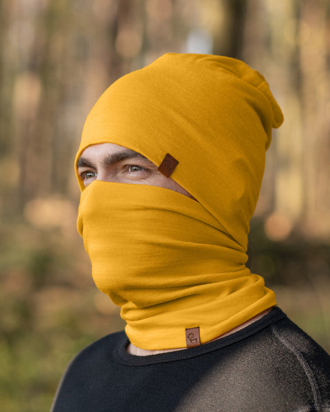 A person with light eyes is wearing a spicy yellow menique beanie that is pulled down to cover part of their forehead and a matching neck gaiter that is pulled up to cover their mouth and nose. They are wearing a dark long-sleeved shirt, and the blurred background suggests an outdoor setting with trees and foliage in muted tones.