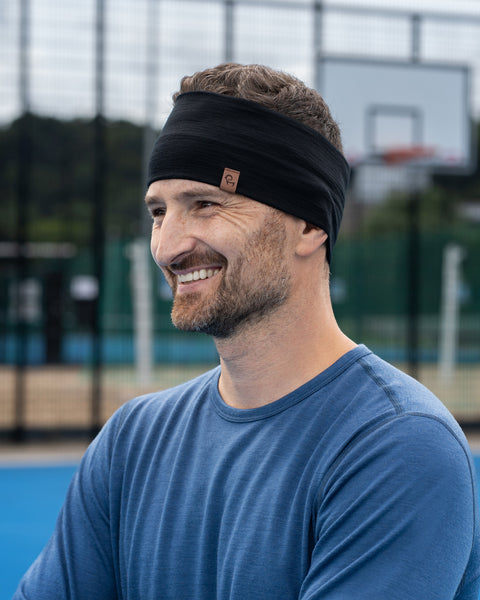A close-up shot of a smiling man looking to the side while standing on a bright blue outdoor sports court. He is wearing a denim-blue long-sleeved shirt and a wide black headband. The background features a blurred chainlink fence and a basketball backboard.