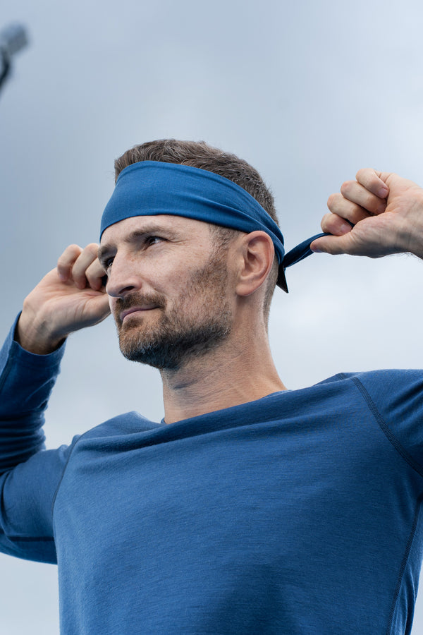 A low-angle shot of a man outdoors tying a blue athletic headband behind his head. He is looking off to the side with a focused expression, wearing a matching blue long-sleeve shirt. The background features a cloudy sky and a blurred, tall stadium light pole.