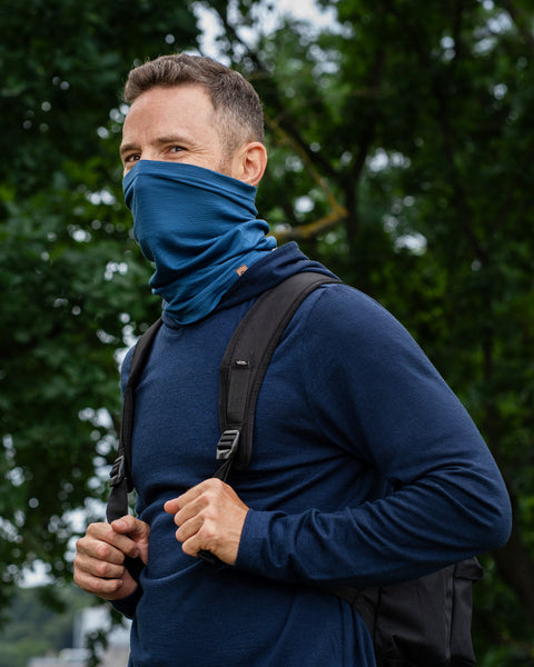 A man outdoors in a wooded area wearing a navy blue long-sleeved hoodie. He has a denim-blue neck gaiter pulled up to cover his lower face and nose. He is grasping the straps of his black backpack and looking back over his shoulder directly at the camera.