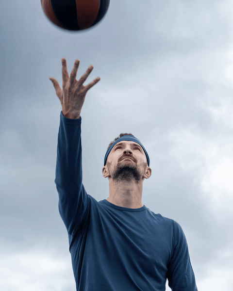 A man in a dark blue long-sleeved shirt and blue headband reaching his hand up toward a basketball against an overcast sky.