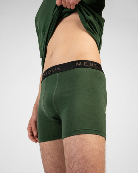 A close-up shot of a man's torso and thighs wearing dark green short boxer briefs with a black logo waistband. He is pulling up his matching dark green t-shirt to reveal his midriff against a light grey background.