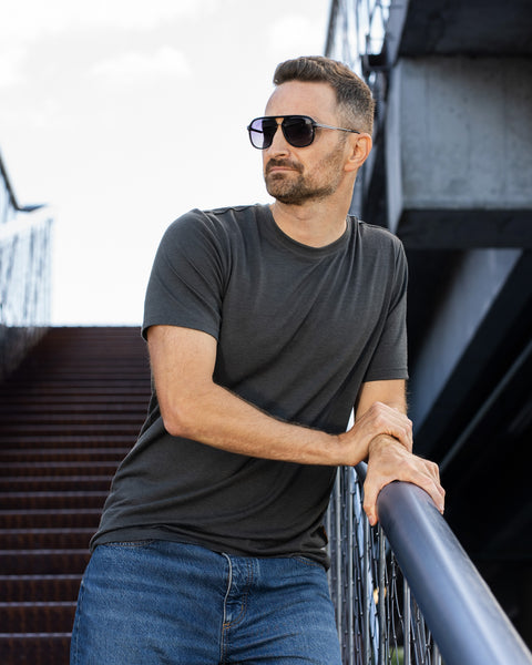 A man with short brown hair and a beard leans against a metal railing next to an outdoor staircase. He is wearing a charcoal grey short-sleeved t-shirt, blue jeans, and dark aviator-style sunglasses, looking off to the left.