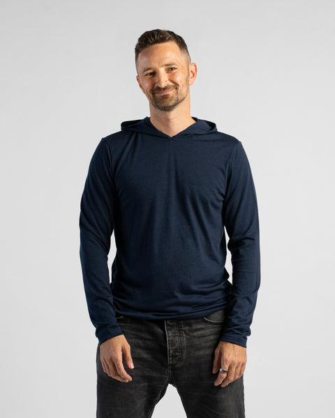 A man wearing a dark navy blue long-sleeve hooded shirt and dark gray jeans, standing against a light gray studio background.