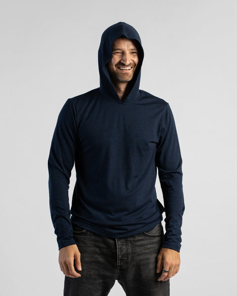 A smiling man with a short beard models a navy blue long-sleeve hoodie with the hood pulled up over his head. He is wearing dark gray jeans and standing against a simple, light gray studio backdrop.