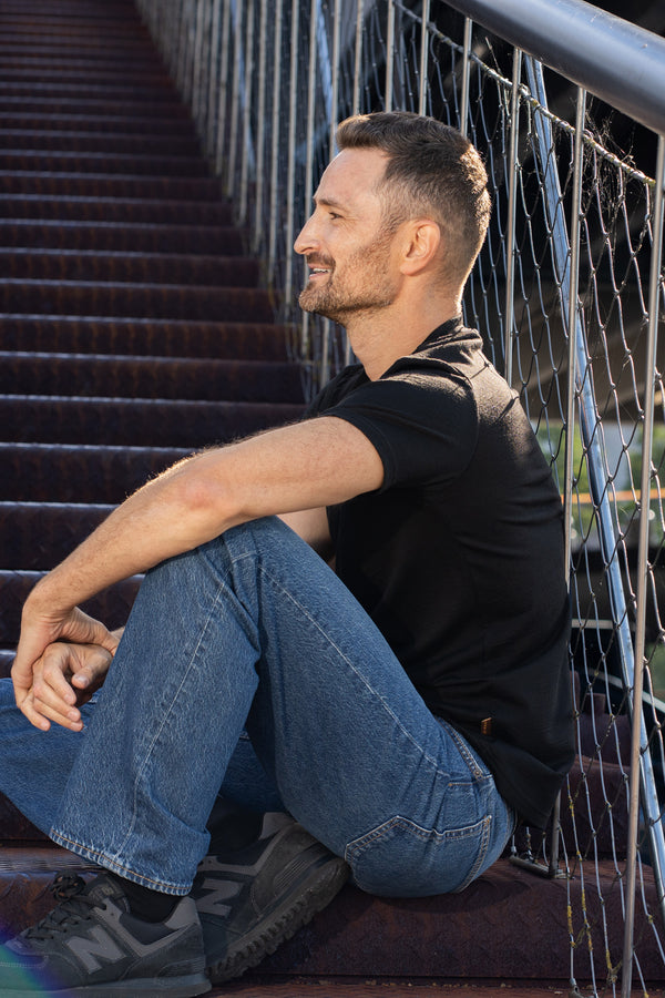 A side profile view of a smiling man sitting on outdoor, rusted steel stairs next to a metal wire railing. He is wearing a black short-sleeved v-neck t-shirt, blue jeans, and dark sneakers, resting his arms comfortably on his knees while looking forward.