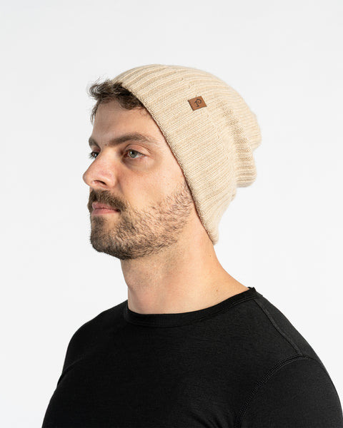 A person with light skin, short dark hair, a mustache, and light-colored eyes is shown in profile against a white background. They are wearing a creamy beige knit beanie with a small brown tag on the side and a black long-sleeved shirt.