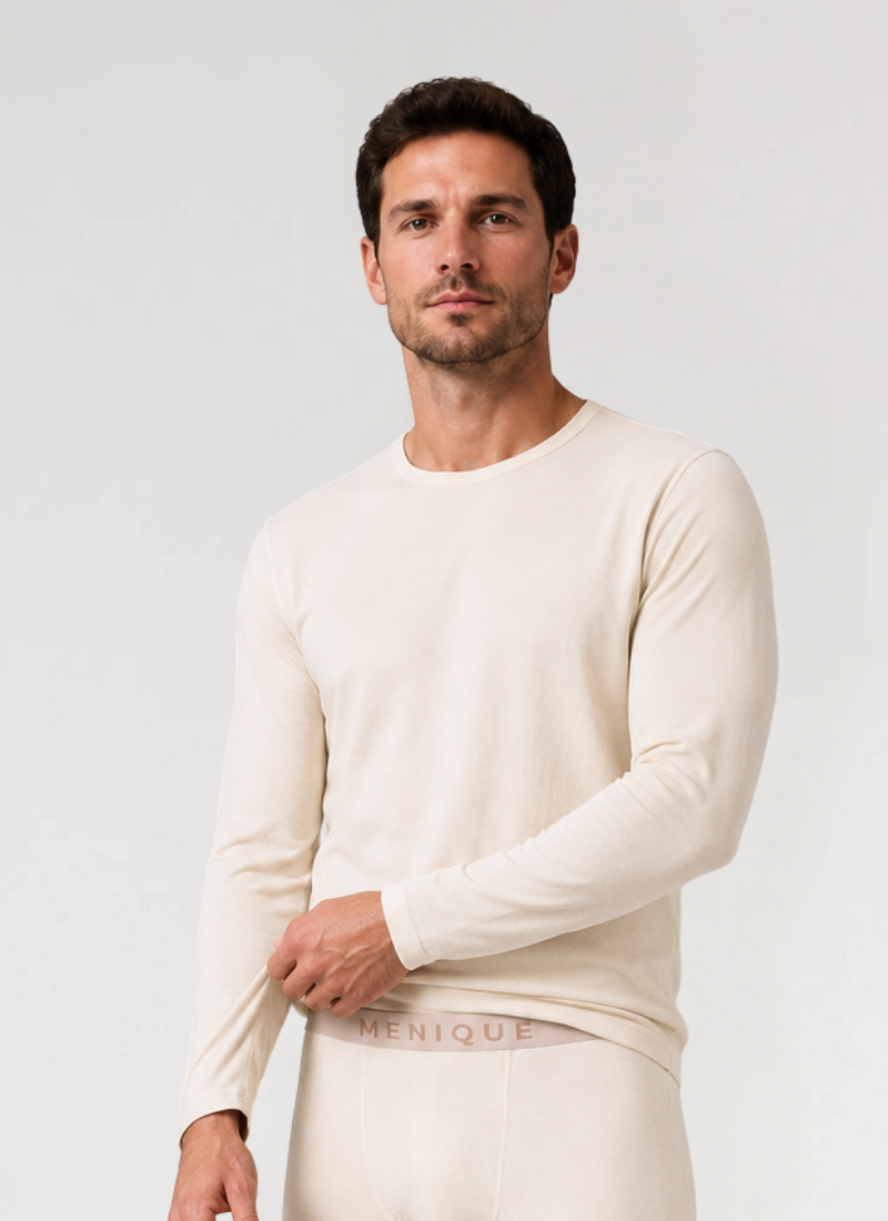 A man standing against a white background wearing a cream-colored long-sleeved base layer top and matching bottoms with a branded waistband, gently pulling the hem of his shirt.