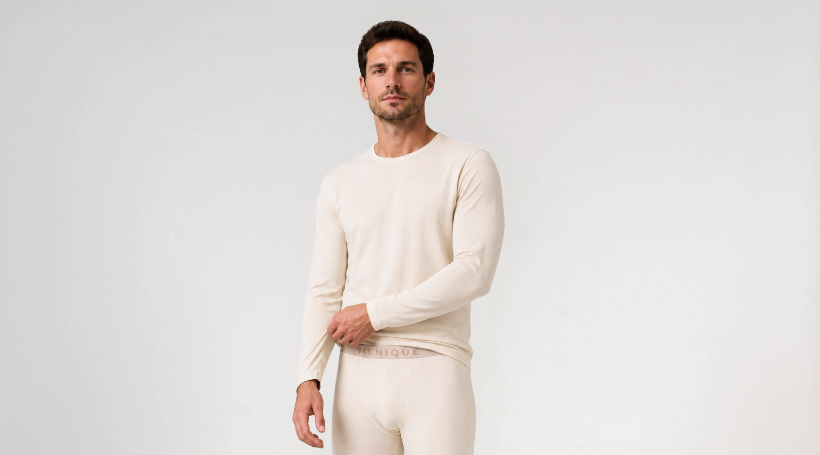 A man standing against a white background wearing a cream-colored long-sleeved base layer top and matching bottoms with a branded waistband, gently pulling the hem of his shirt.