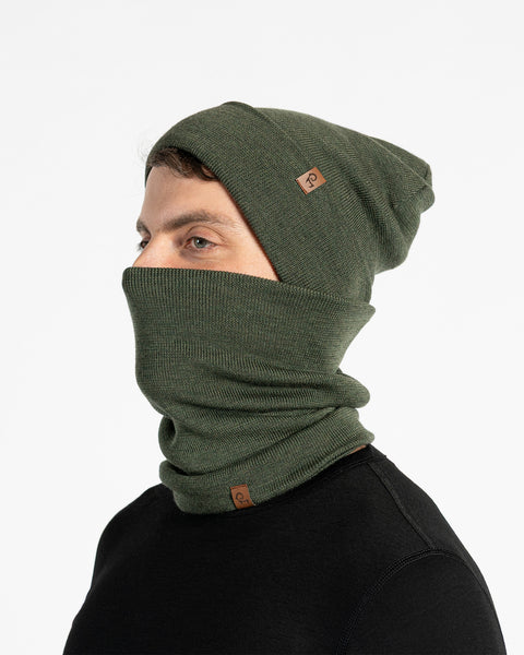 A male model, viewed in profile, wears a dark green beanie and a matching neck gaiter that covers his nose and mouth. Both the beanie and gaiter feature a small, brown leather patch with a Menique logo. He has a short beard and dark hair. The background is solid white.