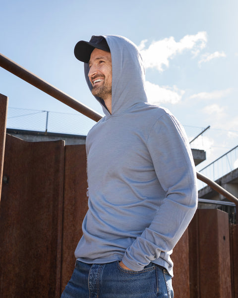 A smiling man standing outdoors near rusted metal barriers, wearing a dusty blue hoodie with the hood pulled up over a black baseball cap. He has one hand in his jeans pocket and is looking to the side.