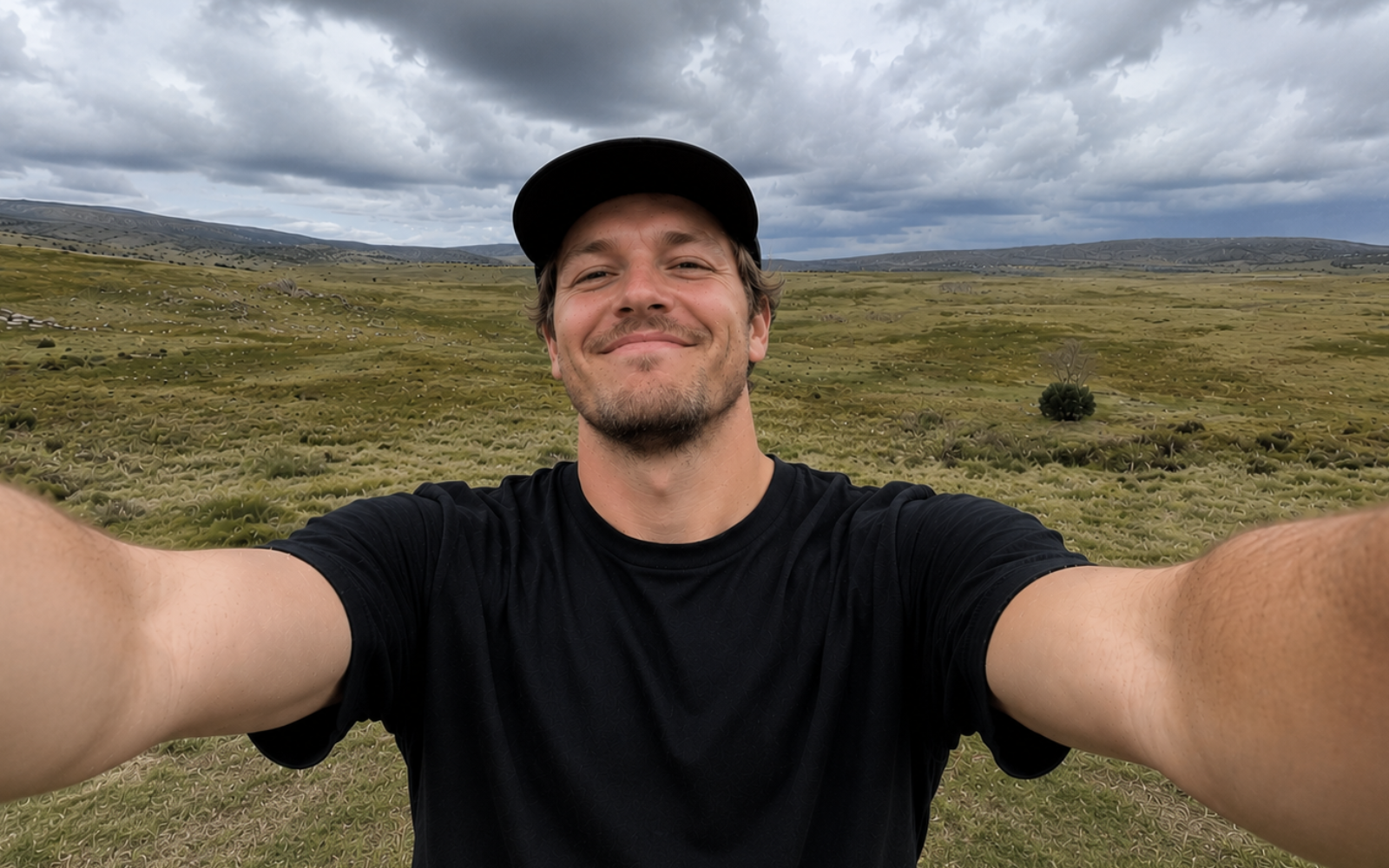 A man wearing a black t-shirt and dark baseball cap taking a smiling selfie in a grassy, hilly landscape under a cloudy sky.