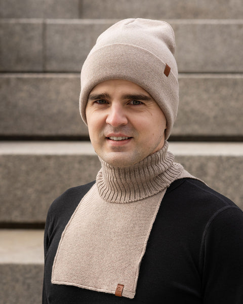 A man smiling slightly and looking directly at the camera. He is wearing a beige knit beanie with a small brown logo patch and a matching beige knit neck warmer with a ribbed collar and flat front panel, worn over a black long-sleeve shirt. The background features blurred concrete stairs.