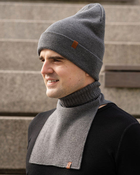 A man shown from the chest up, turned slightly to his right and smiling. He is wearing a dark grey knit beanie with a small brown logo patch and a matching dark grey knit neck warmer over a black long-sleeve shirt. The background consists of blurred concrete stairs.